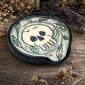 May include: A round ceramic spoon rest with a dark blue rim and a light blue center. The center features a hand-painted cream-colored skull with dark blue eye sockets and nose cavity, surrounded by a decorative pattern of green leaves.