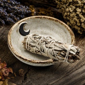 May include: A round, speckled ceramic dish with a dark brown crescent moon accent on the rim. Inside the dish rests a bundle of dried sage leaves tightly wrapped with thin white string, creating a textured, earthy composition.