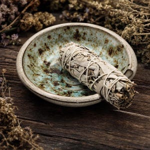 May include: A round ceramic bowl with a speckled light blue and brown glaze containing a bundle of dried white sage leaves wrapped in thin white string. The bowl has a thick rim and a shallow basin, resting in a horizontal position.