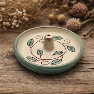 May include: A round ceramic incense holder with a cream-colored base and a teal-tinted rim. The center features a raised conical post for holding incense sticks, surrounded by a hand-painted brown vine pattern with small green leaves.