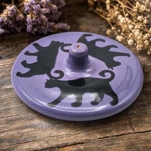 May include: A round, lavender-colored ceramic incense burner featuring a central holder for incense sticks. The surface is decorated with three black cat silhouettes arranged in a circular pattern around the center post, with a glossy finish.