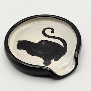 May include: A black and white ceramic spoon rest. The spoon rest has a black rim and a cream-colored interior. A black cat silhouette is painted on the interior. The spoon rest has a curved shape with a small spout.