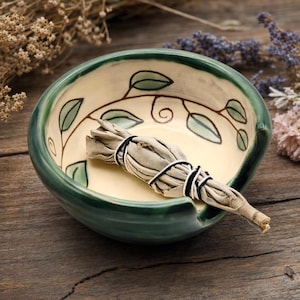 May include: A round ceramic bowl with a dark green rim and a cream-colored interior featuring a hand-painted brown vine design with green leaves. A bundle of dried white sage tied with black and white string rests inside the bowl.
