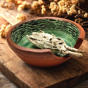 May include: A handcrafted, terracotta-colored ceramic bowl with a green interior and black patterned rim. The bowl holds a bundle of dried sage tied with twine, perfect for smudging rituals. The bowl sits on a wooden surface.