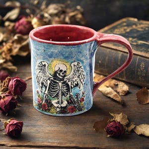 May include: A ceramic mug with a light blue speckled exterior and a red interior. The front features a black and white illustration of a winged skeleton with a yellow halo, surrounded by red roses. The mug has a curved red handle on the right side.