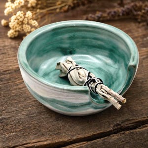 May include: A round ceramic bowl with a glossy teal and white marbled glaze finish. The bowl features a distinct side notch designed for resting items. Inside the bowl lies a bundle of dried white sage leaves tied with black string.