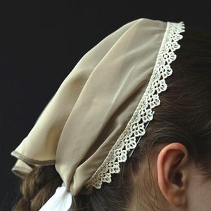 May include: A beige head covering with a white lace trim and a white ribbon tie. The head covering is worn on the head of a person with brown hair. The background is black.