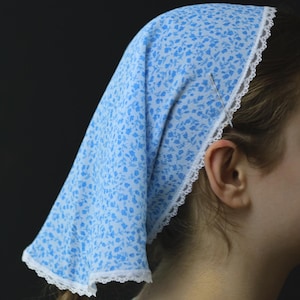 May include: A light blue headscarf with a white floral pattern and white lace trim. The head covering is triangular and worn on the head. The background is black.