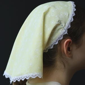 May include: A pale yellow head covering with a white floral pattern and white lace trim. The head covering is worn over the hair, which is styled in a long, brown braid. The background is a dark, solid color.