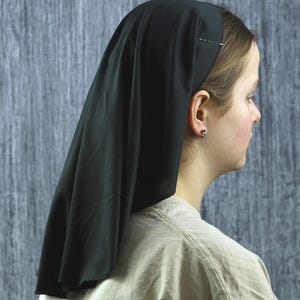 May include: A black head covering, likely a veil or scarf, draped over the head and shoulders. The fabric is dark and appears to be lightweight. A decorative pin secures the veil to the hair. The person is wearing a light-colored shirt.