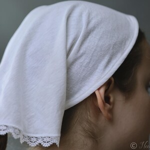 May include: A white head covering with a delicate lace trim. The fabric is draped over the head and tied at the back. The head covering is made of a soft, lightweight material.