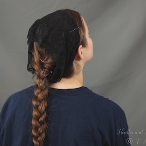 May include: A black lace head covering featuring a delicate floral pattern. The fabric is lightweight and semi-sheer, designed to be worn over the hair. The lace has a scalloped edge detail along the perimeter, providing a textured appearance.