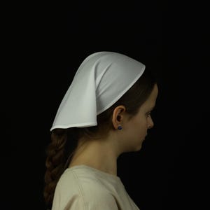 May include: A white head covering, possibly a bonnet or cap, is worn by a person with brown hair. The head covering is made of a smooth, opaque fabric. The person is wearing a light-colored top. The background is black.