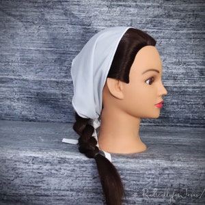 May include: A white cotton head covering with a simple design. The head covering is tied at the back with a white ribbon.