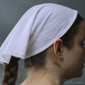 May include: A white head covering, likely a bonnet or scarf, is draped over the head. The fabric is smooth and appears to be cotton or a similar lightweight material. The head covering is worn over the hair, with a braid visible.