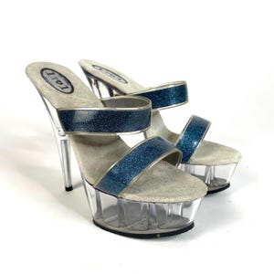 Puede incluir: Par de tacones de plataforma de brillo azul con correas transparentes y una plataforma transparente. Los tacones miden aproximadamente 15 cm de alto y la plataforma unos 5 cm. Los zapatos son de la marca "Lolli".