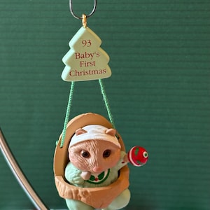 May include: A Christmas ornament featuring a baby raccoon in a cradle. The raccoon wears a white cap and green outfit, holding a red and green ornament. A green tree-shaped tag reads "93 Baby's First Christmas".