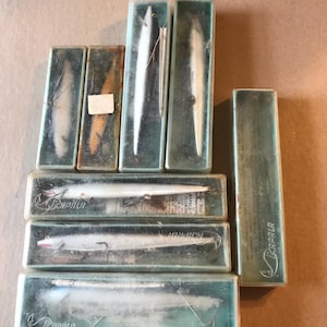 May include: A collection of vintage Rapala fishing lures in their original packaging. The lures are silver and white with black accents and are displayed in clear plastic boxes with green and white labels. The boxes are stacked on a brown surface.
