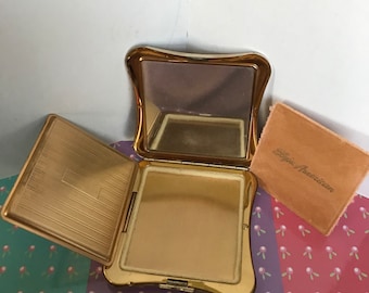 May include: A gold-colored compact mirror with a hinged lid and a small, rectangular mirror inside. The compact is open and the lid is propped up. The compact is sitting on a patterned fabric surface. A small, brown velvet pouch with the text "Lady Americana" is visible in the background.