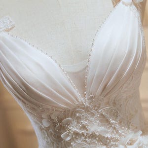 Op de afbeelding: Close-up van een witte trouwjurk lijfje. De jurk heeft een V-halslijn met delicate, met kralen bezette bandjes en versieringen. Bloemenkant en parelaccenten sieren het lijfje en creëren een romantische en elegante look. De stof lijkt een zacht, vloeiend materiaal te zijn.