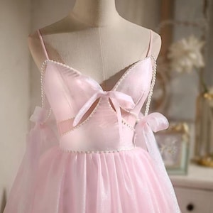Puede incluir: Vestido de fiesta rosa claro sobre un maniquí. El vestido presenta tirantes finos, un corpiño adornado con lazo y una falda amplia. Delicadas perlas adornan el escote y el corpiño. El vestido está hecho de una tela transparente y brillante.