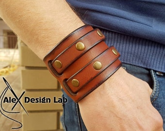 May include: A brown leather cuff bracelet with two straps and brass studs. The bracelet is worn on a person's wrist.
