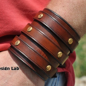 May include: A brown leather bracelet with multiple layers and brass accents. The bracelet is worn on a person's wrist.