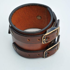 May include: A wide brown leather cuff bracelet featuring two horizontal straps with dark metal buckles and matching rivets. The leather has a smooth, polished finish with darkened edges, and the interior shows the natural, textured brown surface.