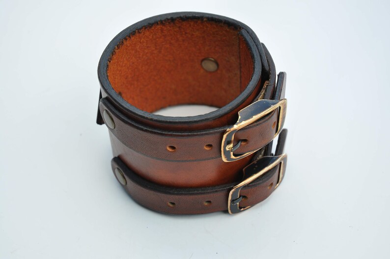 May include: A wide brown leather cuff bracelet featuring two horizontal straps with dark metal buckles and matching rivets. The leather has a smooth, polished finish with darkened edges, and the interior shows the natural, textured brown surface.