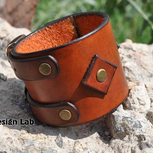 May include: A wide brown leather cuff bracelet featuring a smooth finish, dark brown edges, and three circular brass-toned metal studs. A diamond-shaped leather patch with a central brass stud is attached to the side. The text Alex Design Lab is visible.
