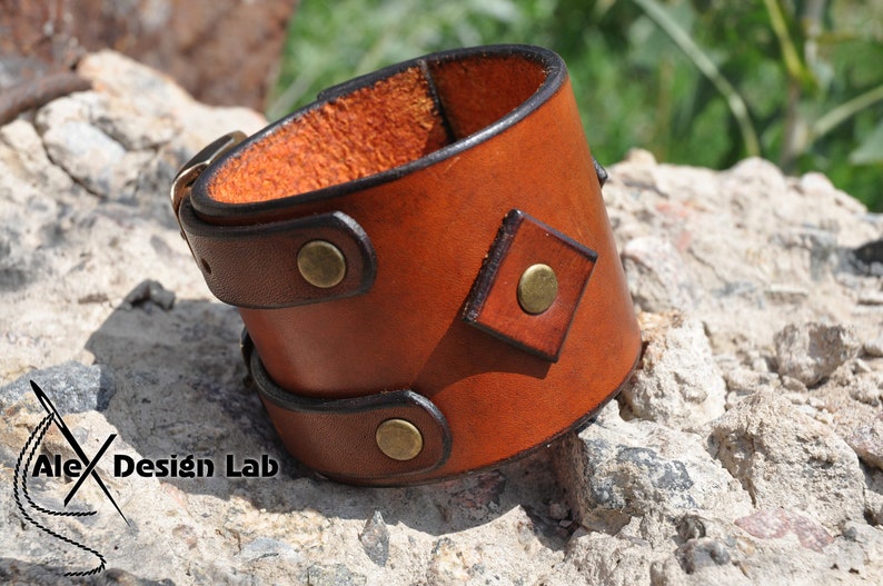 May include: A wide brown leather cuff bracelet featuring a smooth finish, dark brown edges, and three circular brass-toned metal studs. A diamond-shaped leather patch with a central brass stud is attached to the side. The text Alex Design Lab is visible.