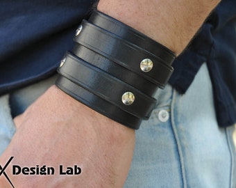 Puede incluir: Un brazalete de cuero negro con tachuelas plateadas. El brazalete es de doble capa y tiene un diseño ancho y plano.
