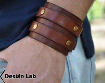Puede incluir: Un brazalete de cuero marrón con tres correas y detalles metálicos de color dorado. El brazalete se lleva en la muñeca.