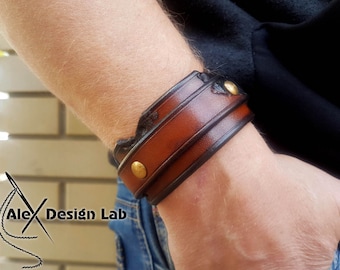 May include: A brown leather bracelet with a black leather trim and gold metal accents. The bracelet is worn on a person's wrist.
