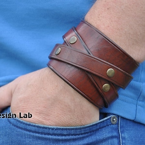May include: A brown leather cuff bracelet with brass accents. The bracelet is worn on a person's wrist.