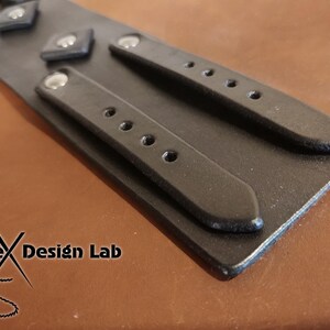 May include: A black leather cuff bracelet featuring two parallel straps with silver-toned metal rivets and multiple adjustment holes. The surface is smooth and matte. The logo Alex Design Lab is printed in black on the lower left corner of the item.