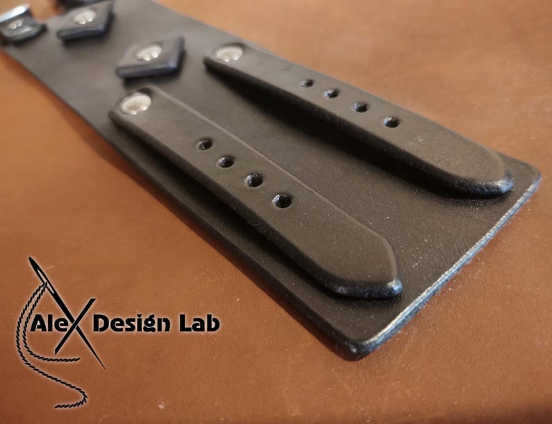 May include: A black leather cuff bracelet featuring two parallel straps with silver-toned metal rivets and multiple adjustment holes. The surface is smooth and matte. The logo Alex Design Lab is printed in black on the lower left corner of the item.