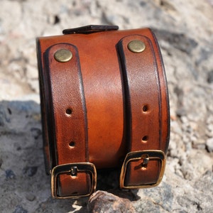 May include: A wide brown leather cuff bracelet featuring two vertical straps with brass-toned metal buckles and rivets. The leather has a smooth, polished finish with darker edges and visible stitching along the length of the straps.