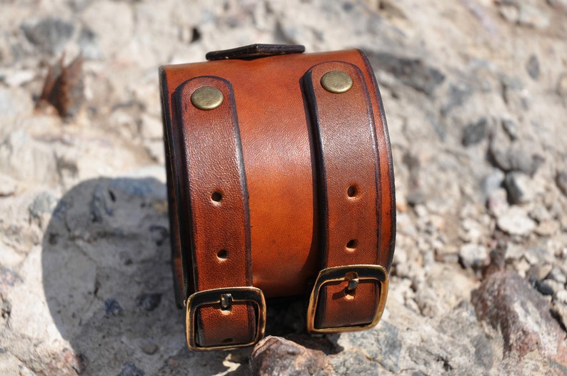 May include: A wide brown leather cuff bracelet featuring two vertical straps with brass-toned metal buckles and rivets. The leather has a smooth, polished finish with darker edges and visible stitching along the length of the straps.