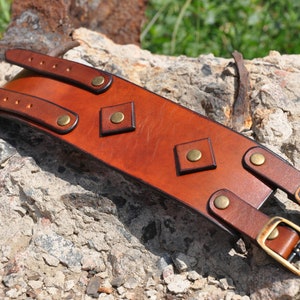 May include: A wide tan leather cuff bracelet featuring two adjustable straps with brass-toned metal buckles and rivets. The center of the cuff is decorated with two diamond-shaped leather patches, each secured with a central brass-toned metal rivet.