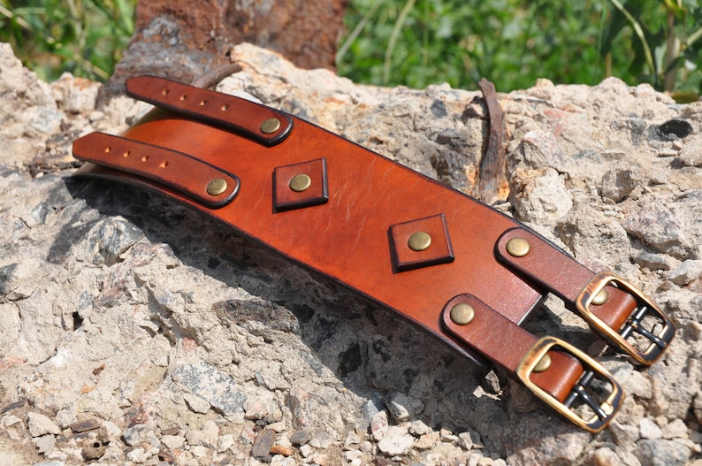 May include: A wide tan leather cuff bracelet featuring two adjustable straps with brass-toned metal buckles and rivets. The center of the cuff is decorated with two diamond-shaped leather patches, each secured with a central brass-toned metal rivet.