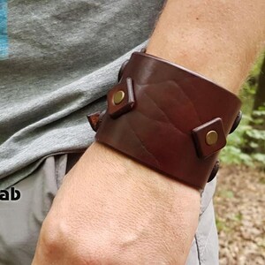May include: A wide dark brown leather cuff bracelet featuring a textured surface and two square leather accents secured with brass-colored metal rivets. The bracelet is worn on the wrist and displays a smooth, polished finish across the leather band.