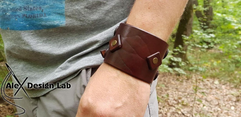 May include: A wide dark brown leather cuff bracelet featuring a textured surface and two square leather accents secured with brass-colored metal rivets. The bracelet is worn on the wrist and displays a smooth, polished finish across the leather band.