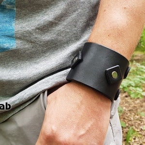 May include: A wide black leather cuff bracelet featuring square leather accents and silver-toned metal snap fasteners. The cuff is worn on the wrist and displays a smooth, matte finish with visible stitching details along the edges of the square tabs.