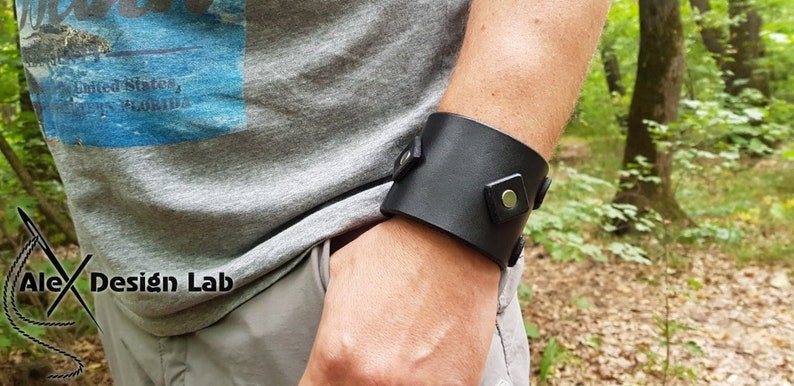 May include: A wide black leather cuff bracelet featuring square leather accents and silver-toned metal snap fasteners. The cuff is worn on the wrist and displays a smooth, matte finish with visible stitching details along the edges of the square tabs.