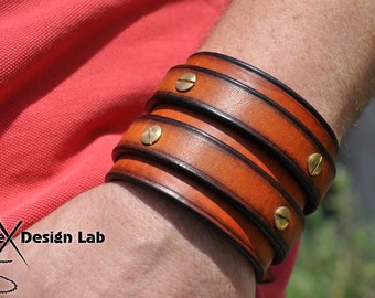 Puede incluir: Un brazalete de cuero de triple vuelta con una correa de cuero marrón y costuras negras. El brazalete tiene detalles y tornillos de latón.