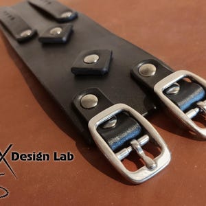 May include: A wide black leather cuff bracelet featuring two silver-toned metal buckles and matching rivets. The surface is adorned with small square leather accents secured by silver studs. The text Alex Design Lab is visible in the corner.