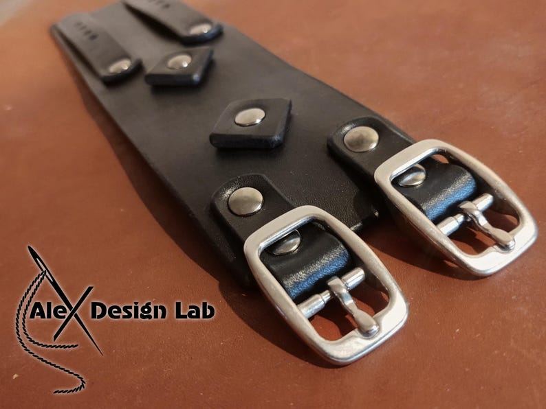 May include: A wide black leather cuff bracelet featuring two silver-toned metal buckles and matching rivets. The surface is adorned with small square leather accents secured by silver studs. The text Alex Design Lab is visible in the corner.