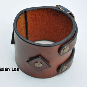 May include: A wide brown leather cuff bracelet featuring a smooth finish, a diamond-shaped leather accent, and three circular brass-toned metal rivets. The interior shows the natural tan texture of the leather. The text Alex Design Lab is visible.