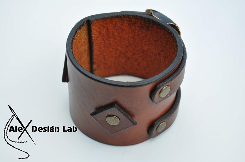 May include: A wide brown leather cuff bracelet featuring a smooth finish, a diamond-shaped leather accent, and three circular brass-toned metal rivets. The interior shows the natural tan texture of the leather. The text Alex Design Lab is visible.